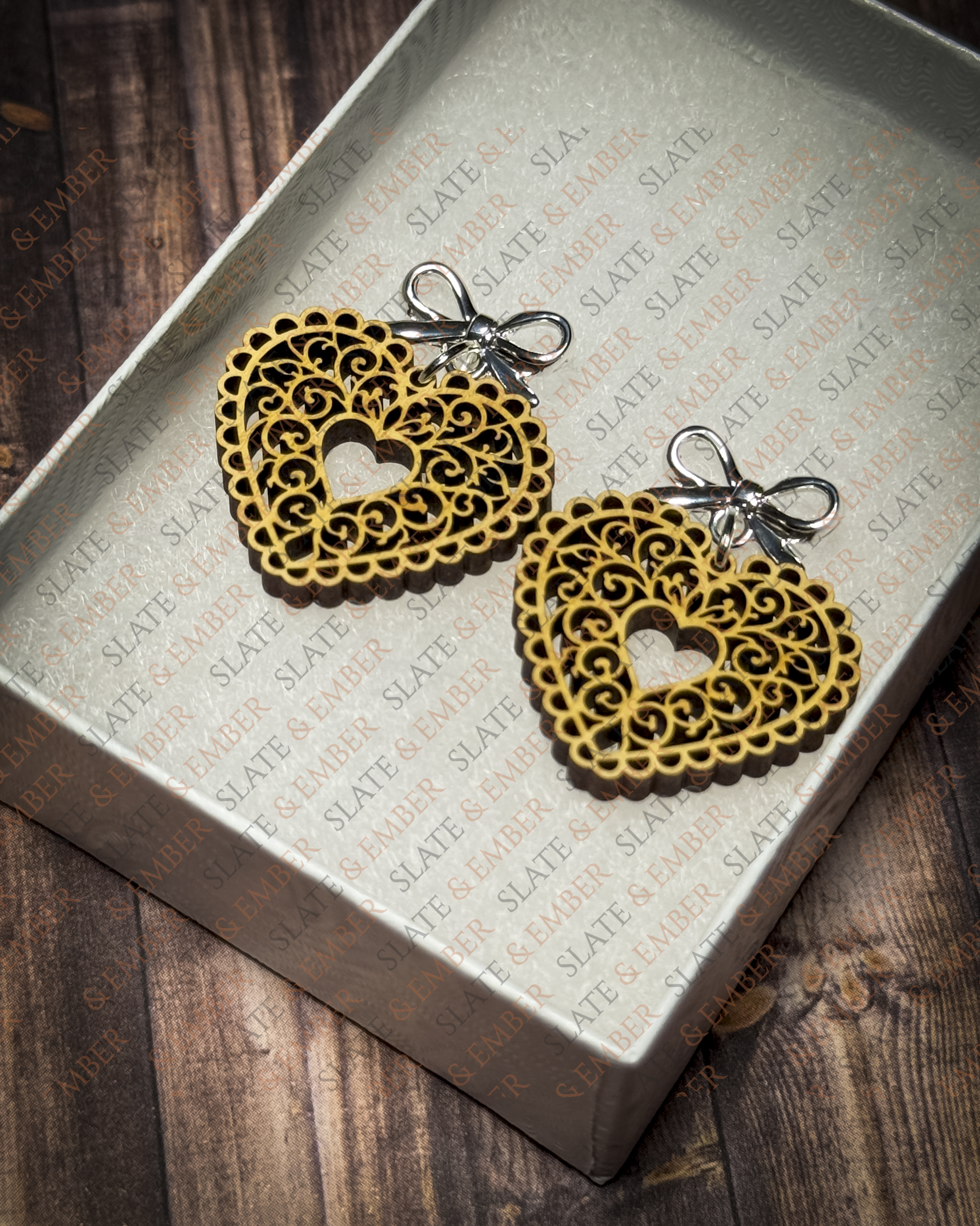 Laser-cut wooden filigree heart earrings with open scrollwork and silver bow stud tops displayed inside a white Slate & Ember jewelry gift box.