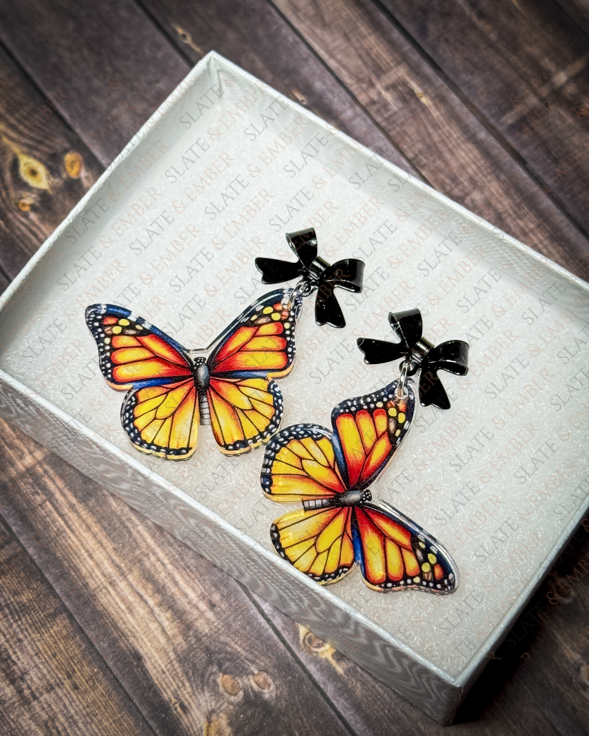 Orange monarch butterfly acrylic earrings with black bow stud tops displayed inside a premium white Slate & Ember jewelry gift box.