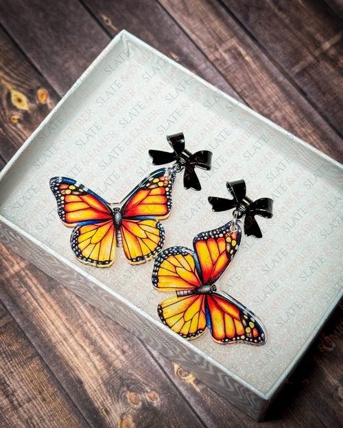 Orange monarch butterfly acrylic earrings with black bow stud tops displayed inside a premium white Slate & Ember jewelry gift box.
