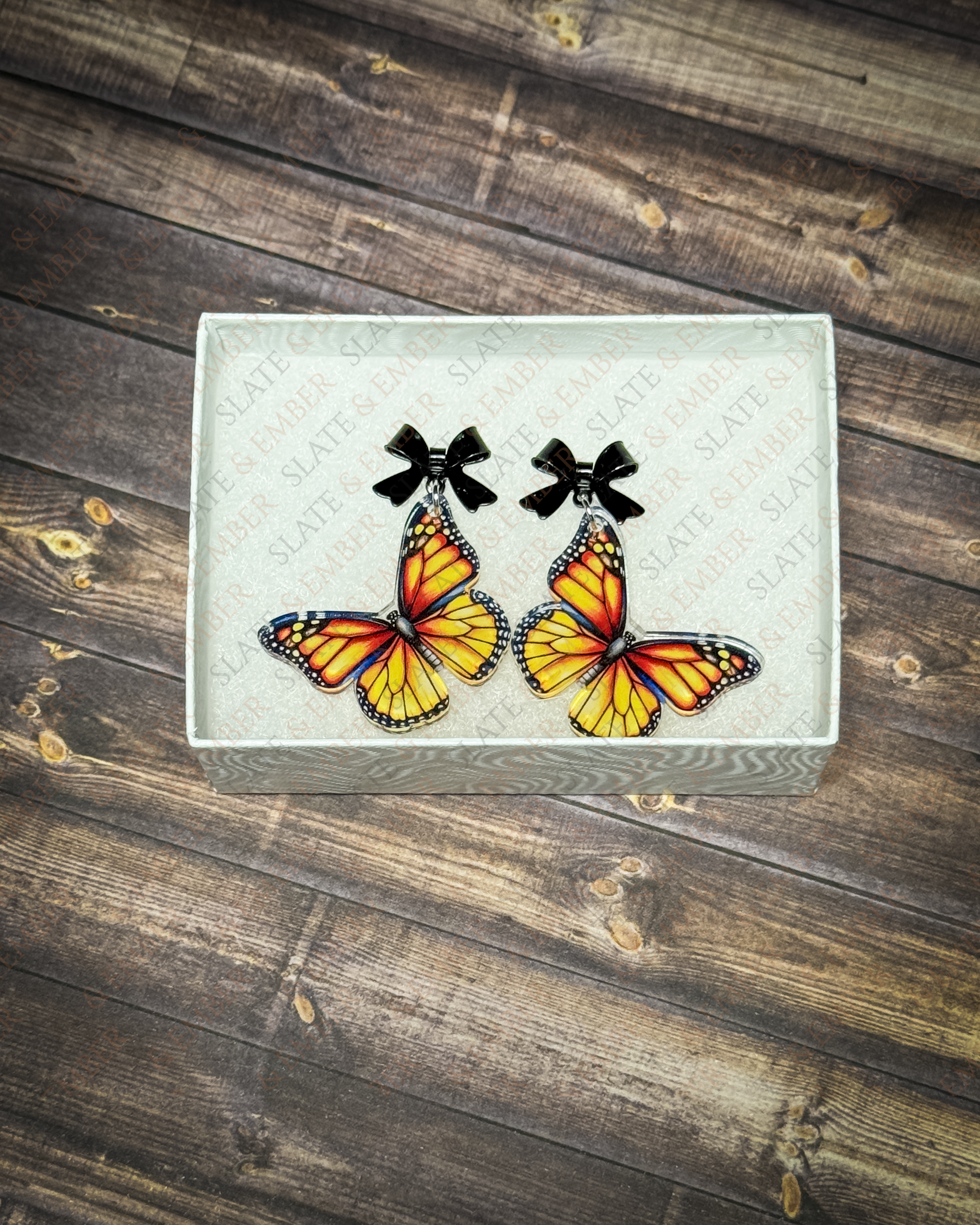 Orange monarch butterfly acrylic earrings with black bow stud tops displayed inside a premium white Slate & Ember jewelry gift box.
