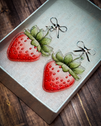 Watercolor-style red strawberry acrylic earrings with green leaves and silver bow stud tops displayed inside a white Slate & Ember jewelry gift box.