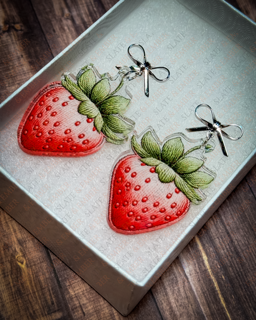 Watercolor-style red strawberry acrylic earrings with green leaves and silver bow stud tops displayed inside a white Slate & Ember jewelry gift box.