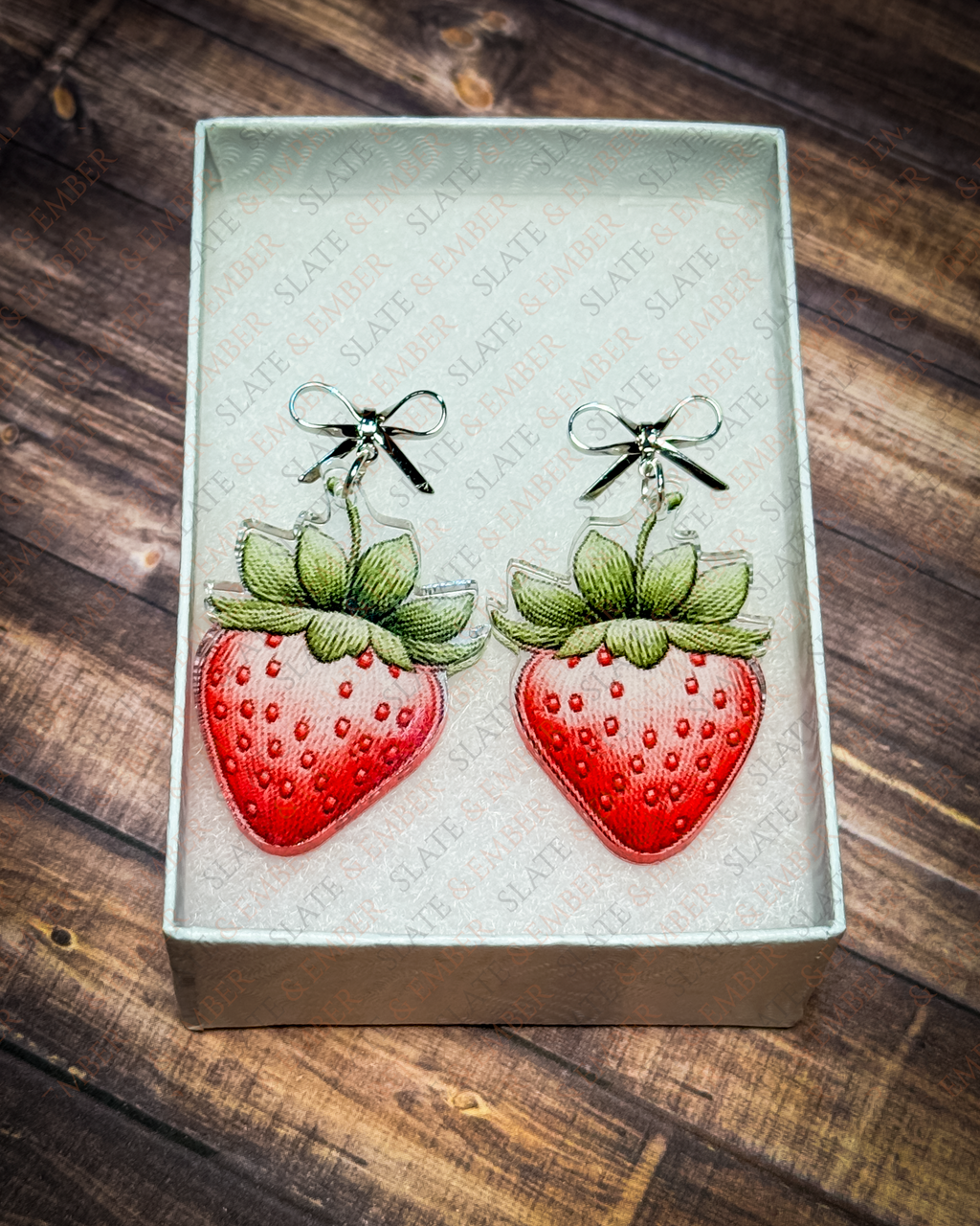 Watercolor-style red strawberry acrylic earrings with green leaves and silver bow stud tops displayed inside a white Slate & Ember jewelry gift box.