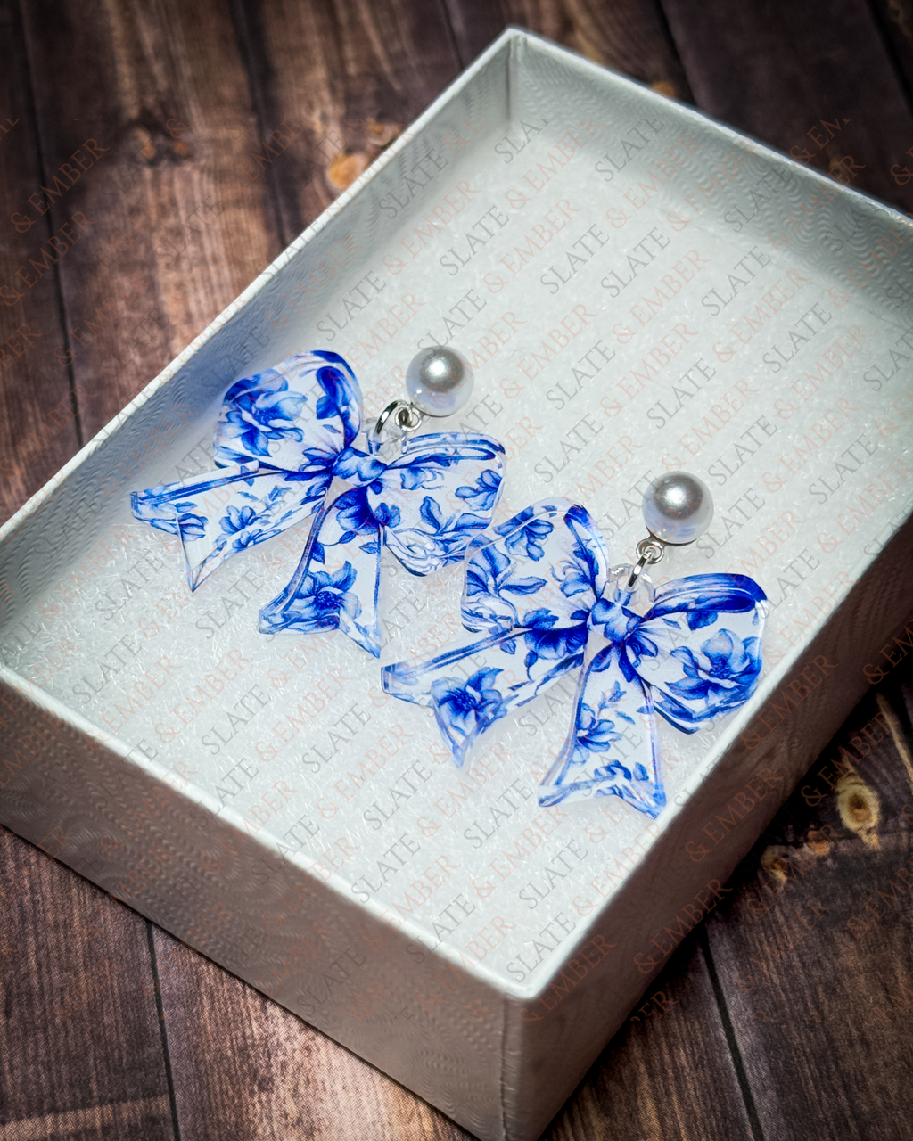 Blue floral chinoiserie acrylic bow earrings with pearl stud tops displayed in a premium white Slate & Ember jewelry gift box.