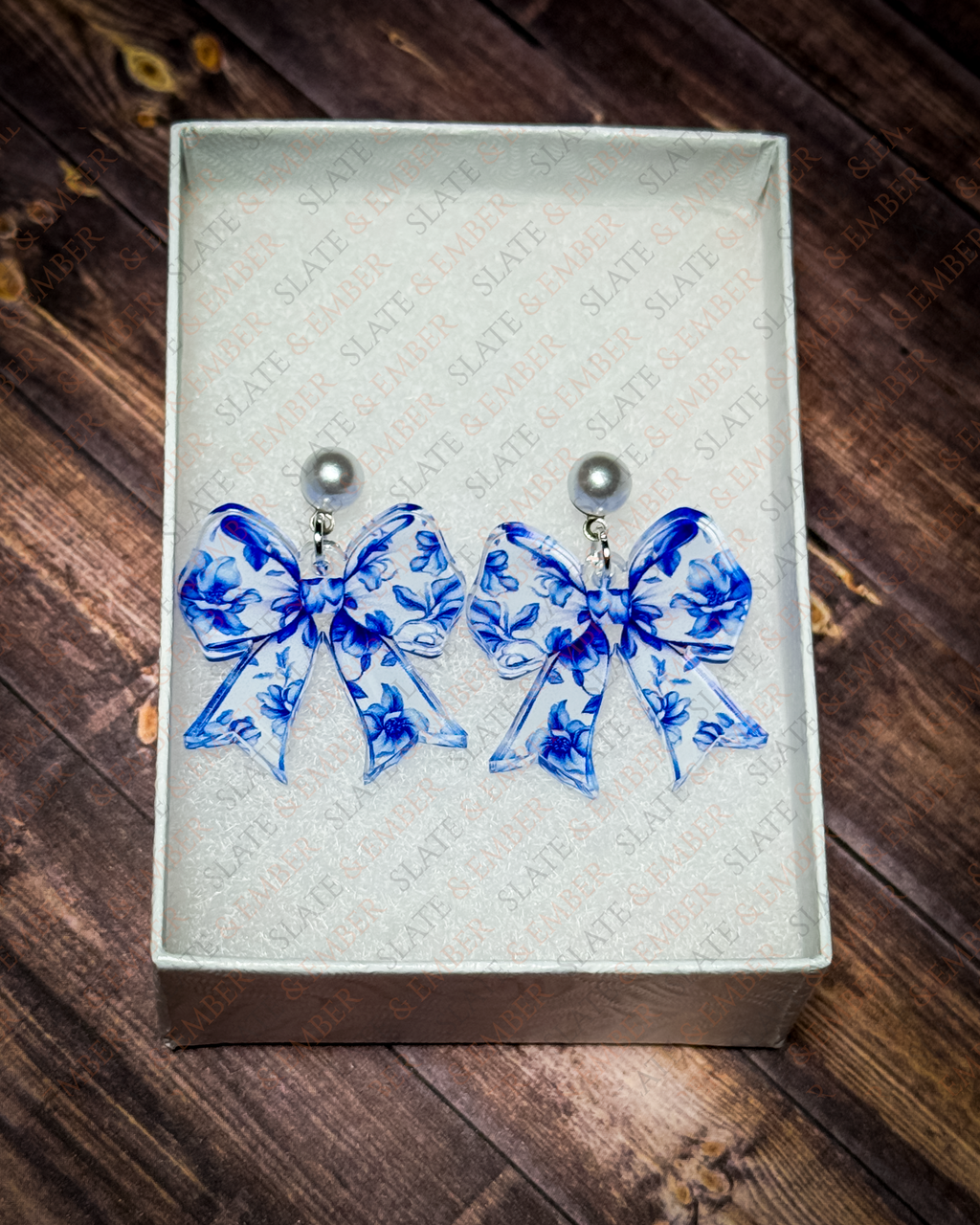 Blue floral chinoiserie acrylic bow earrings with pearl stud tops displayed in a premium white Slate & Ember jewelry gift box.