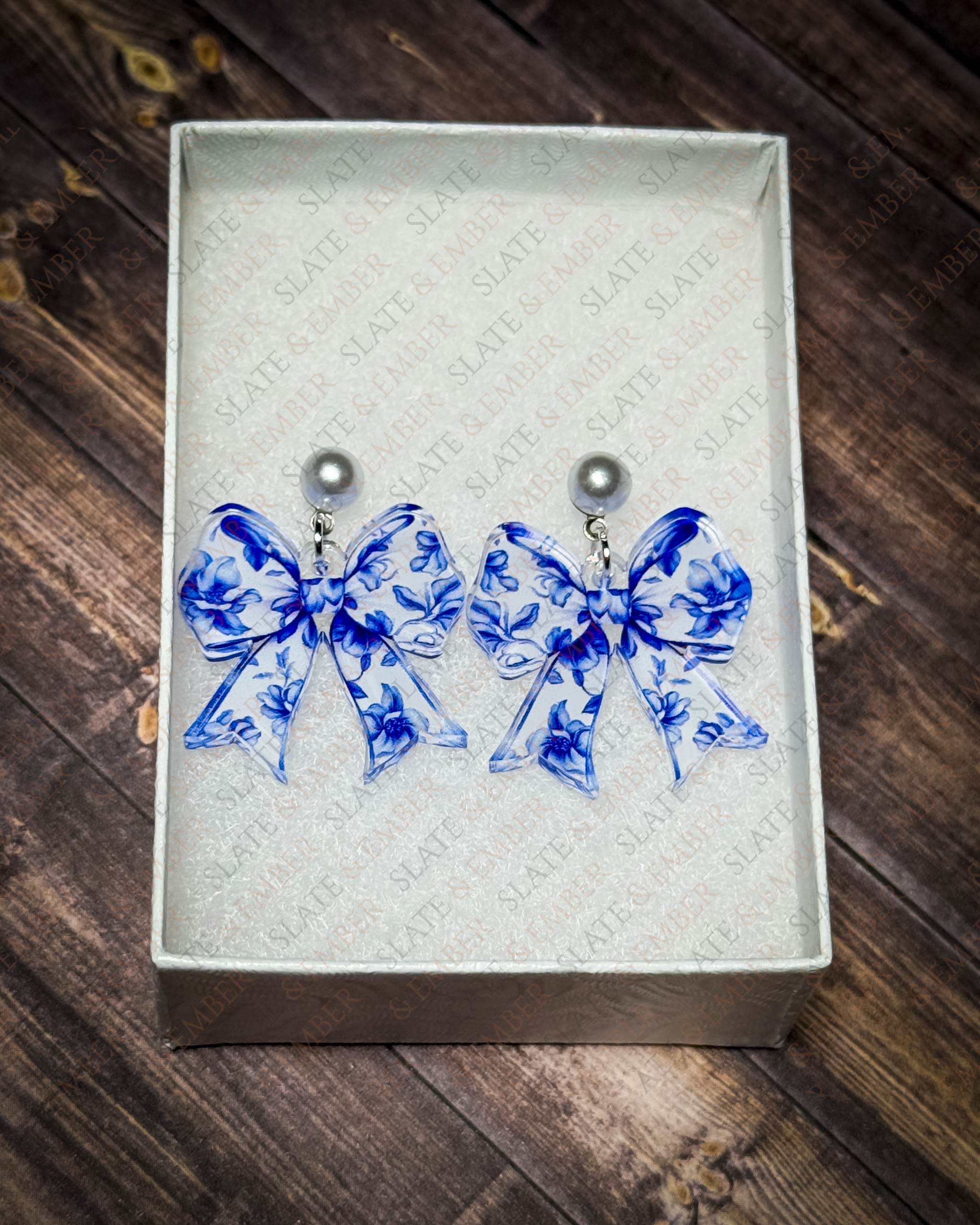 Blue floral chinoiserie acrylic bow earrings with pearl stud tops displayed in a premium white Slate & Ember jewelry gift box.