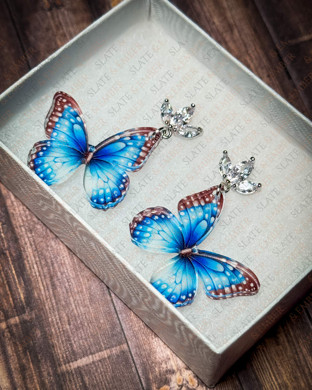 Blue butterfly acrylic earrings with crystal leaf stud tops displayed inside a premium white Slate & Ember jewelry gift box.