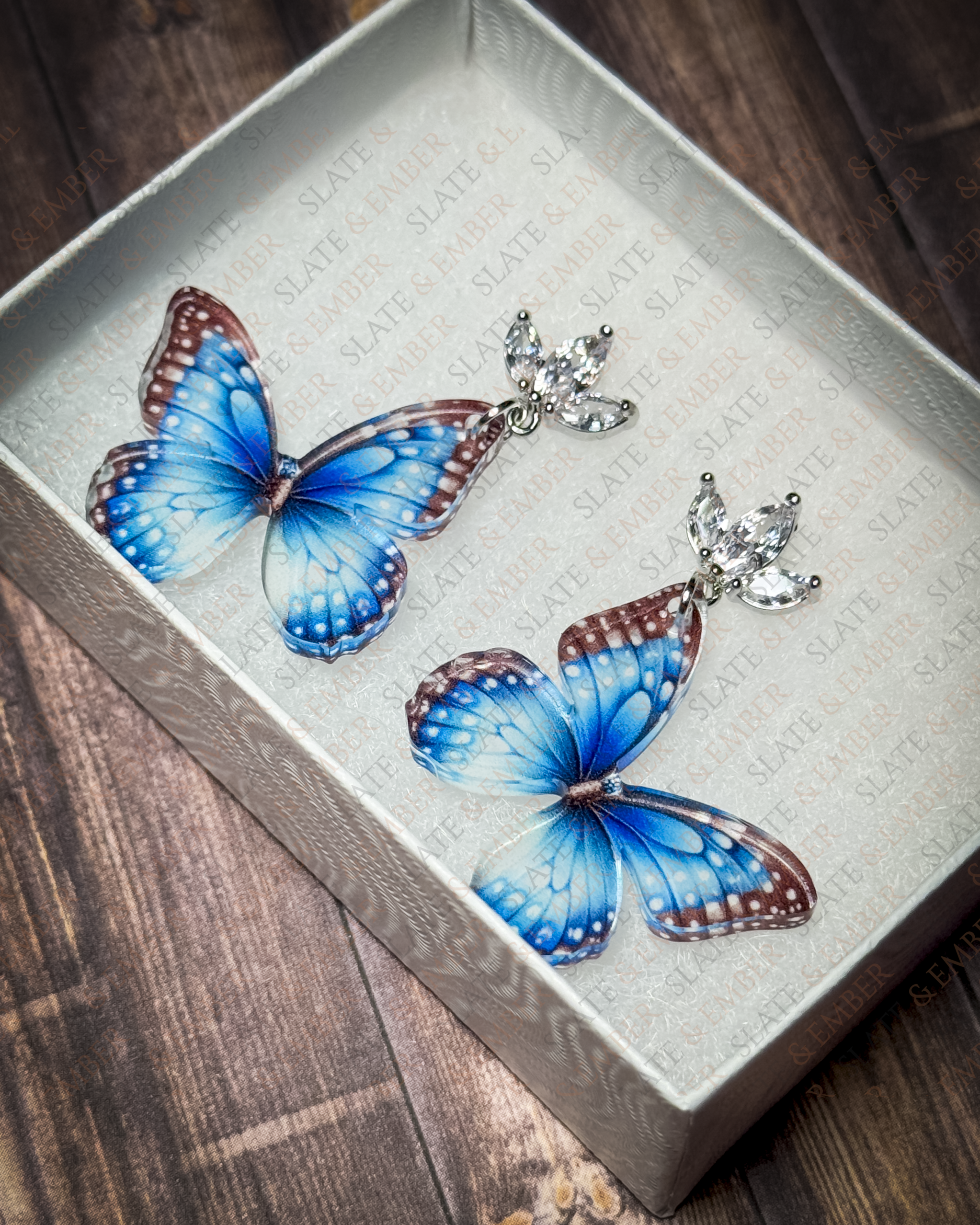 Blue butterfly acrylic earrings with crystal leaf stud tops displayed inside a premium white Slate & Ember jewelry gift box.