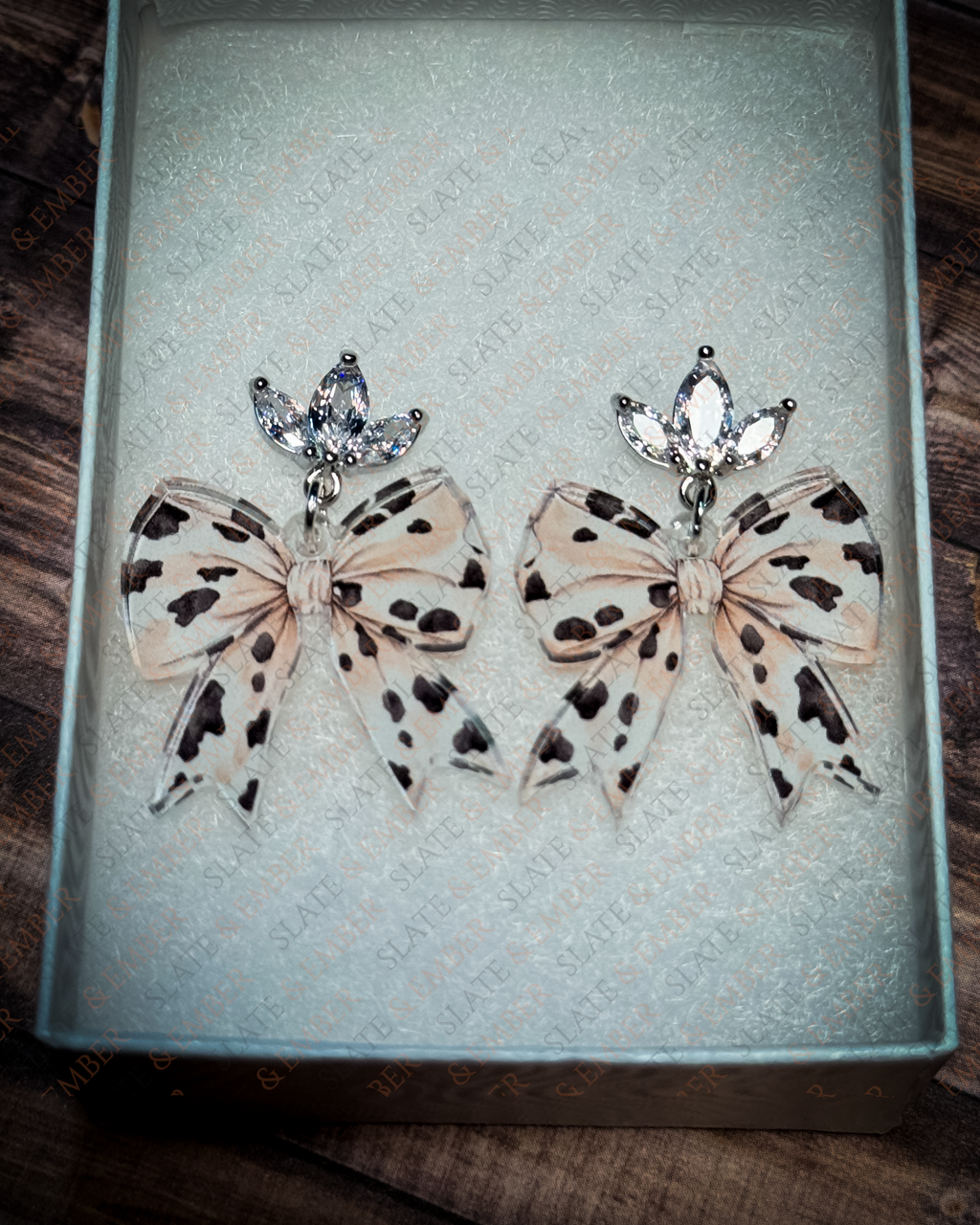 Neutral cow print acrylic bow earrings with crystal leaf stud tops displayed inside a premium white Slate & Ember jewelry gift box.