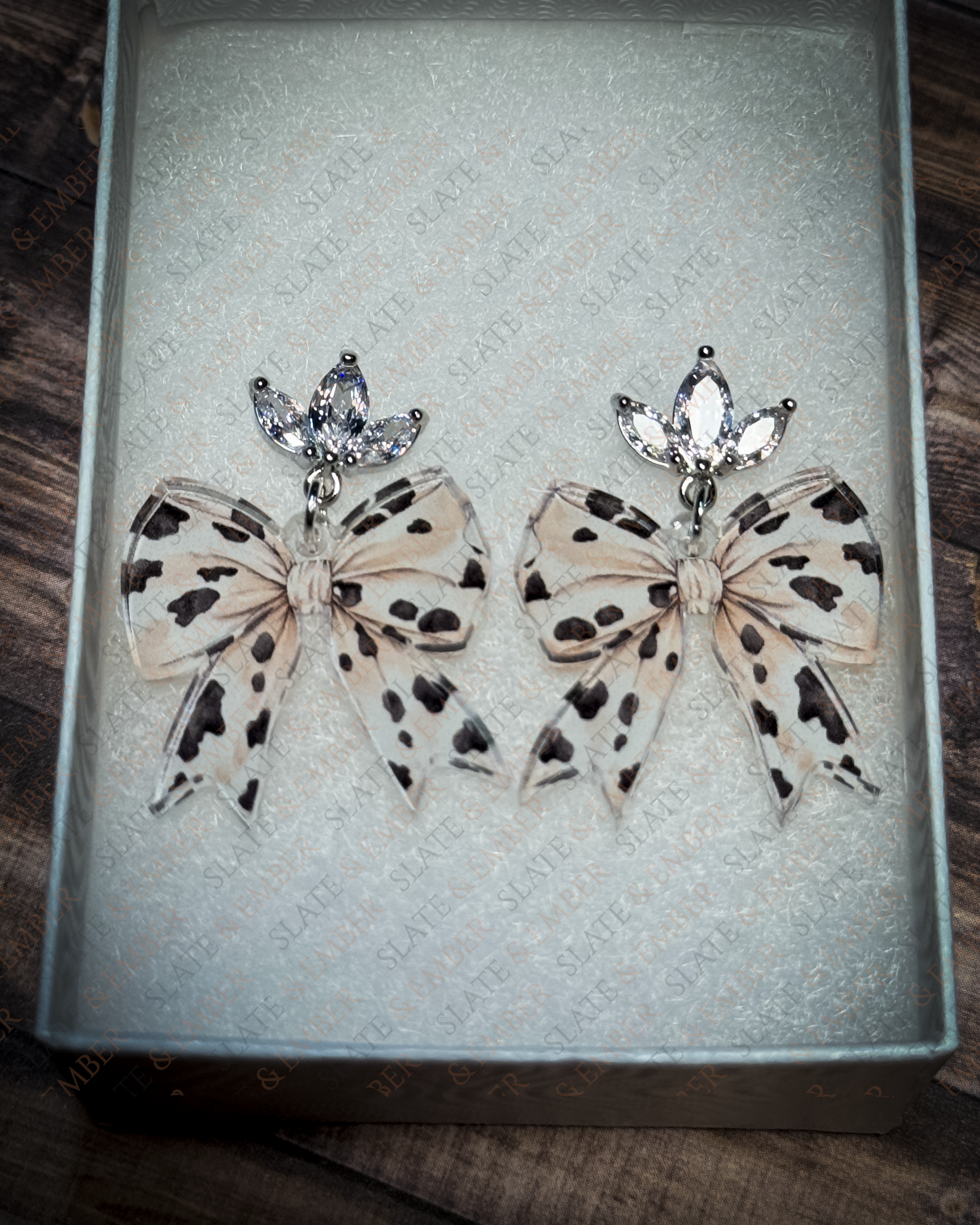 Neutral cow print acrylic bow earrings with crystal leaf stud tops displayed inside a premium white Slate & Ember jewelry gift box.