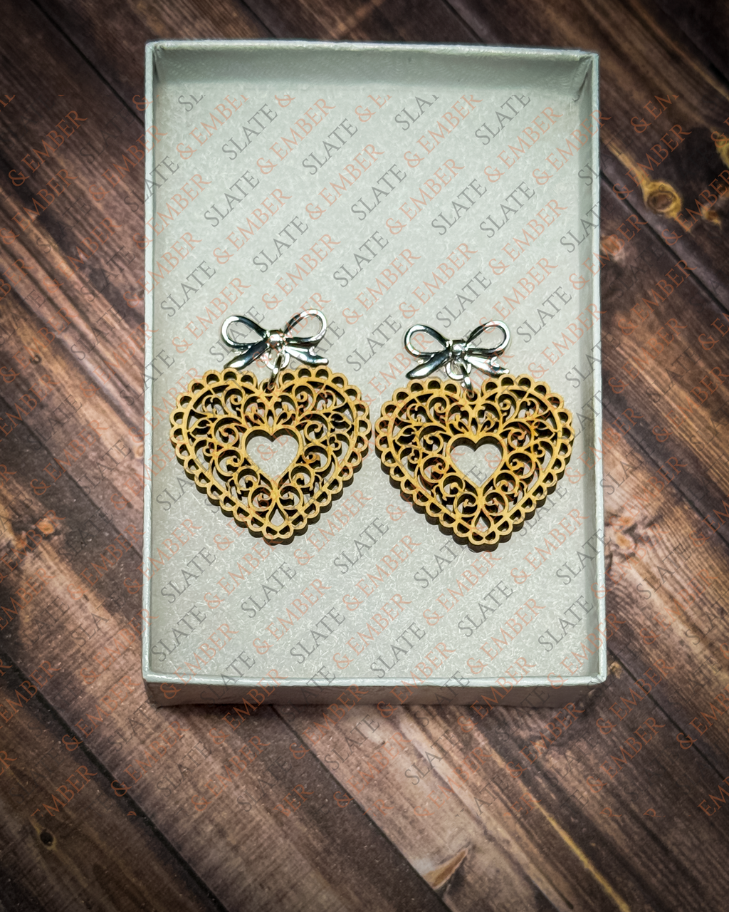 Laser-cut wooden filigree heart earrings with open scrollwork and silver bow stud tops displayed inside a white Slate & Ember jewelry gift box.