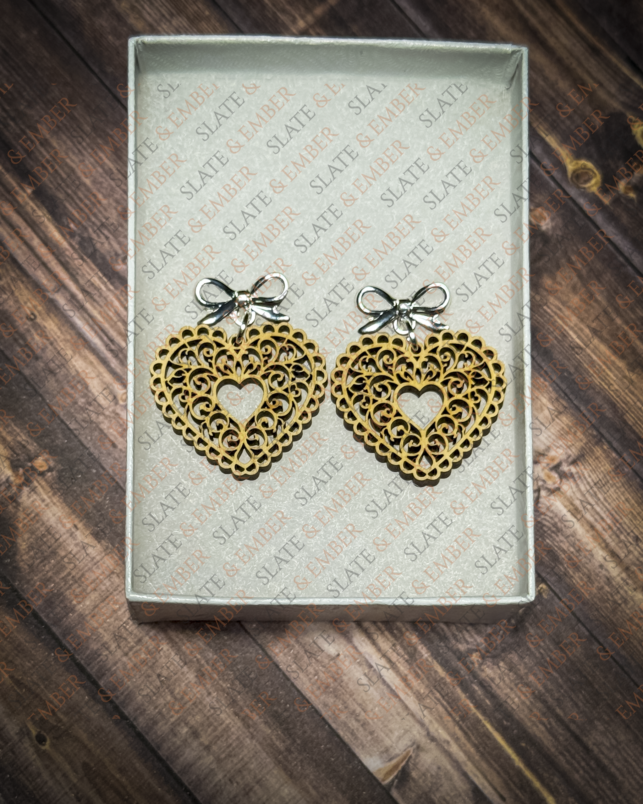 Laser-cut wooden filigree heart earrings with open scrollwork and silver bow stud tops displayed inside a white Slate & Ember jewelry gift box.