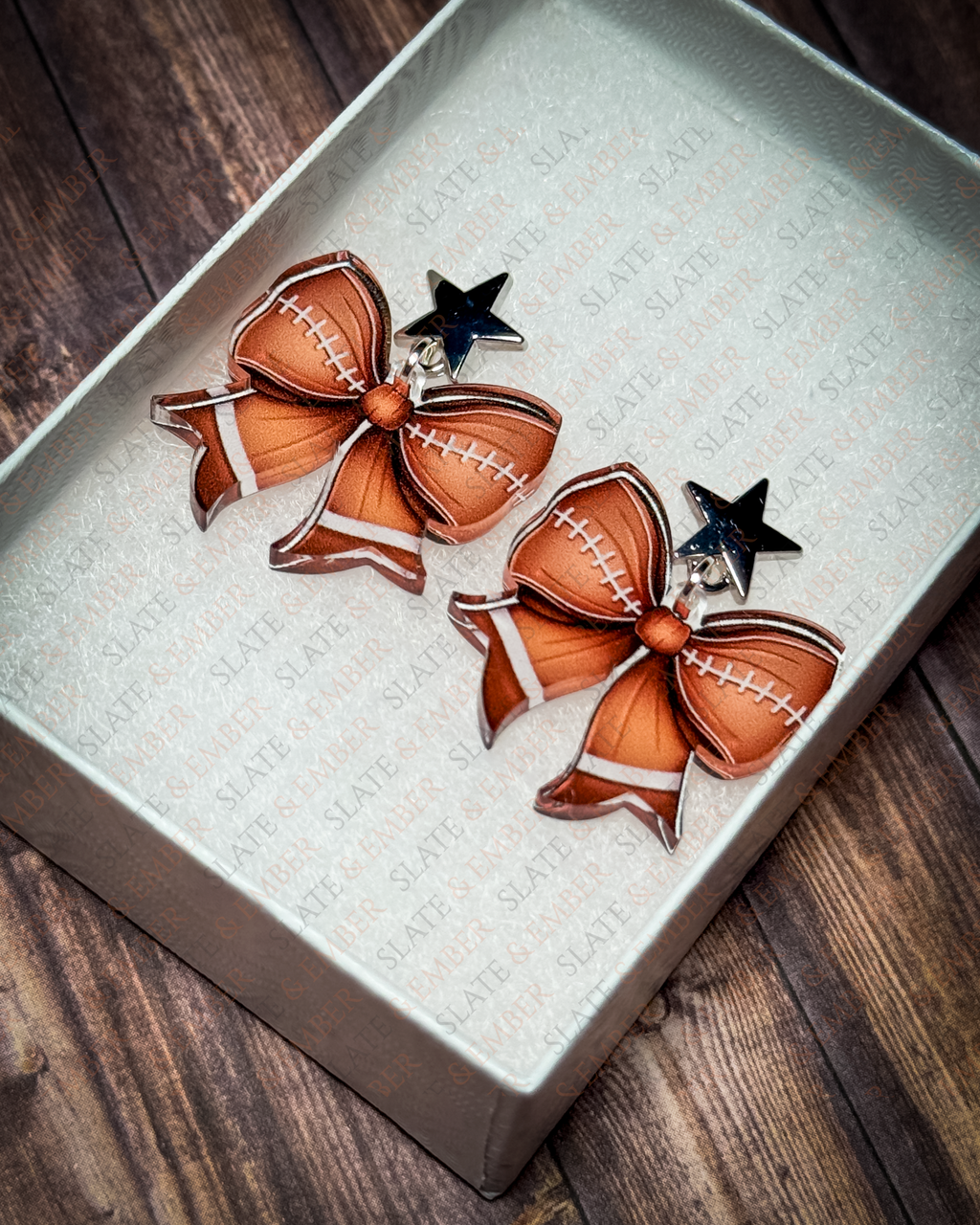 Football bow acrylic earrings with silver star stud tops displayed inside a premium white Slate & Ember jewelry gift box.