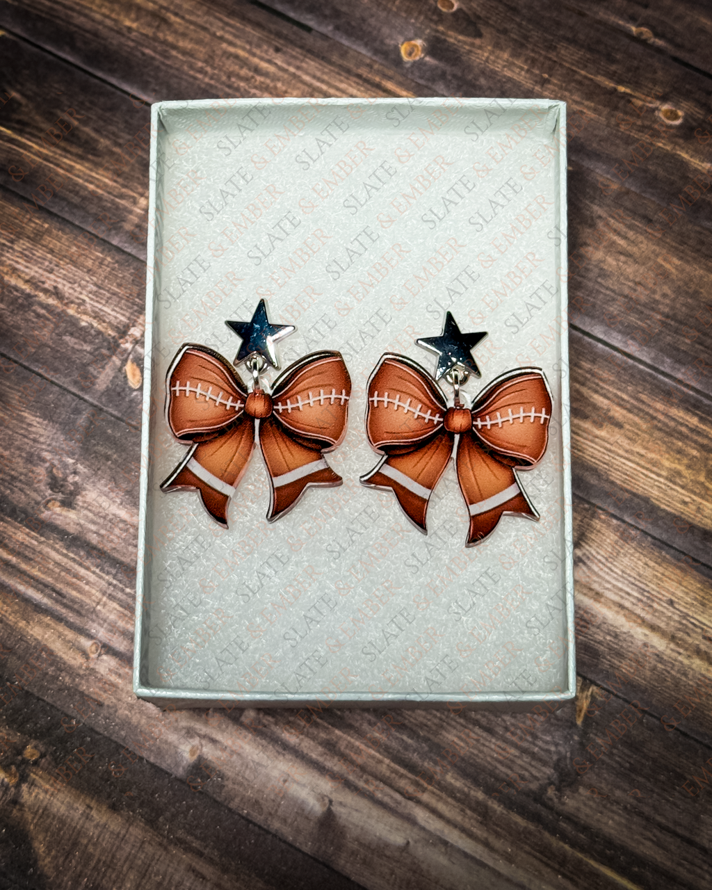 Football bow acrylic earrings with silver star stud tops displayed inside a premium white Slate & Ember jewelry gift box.