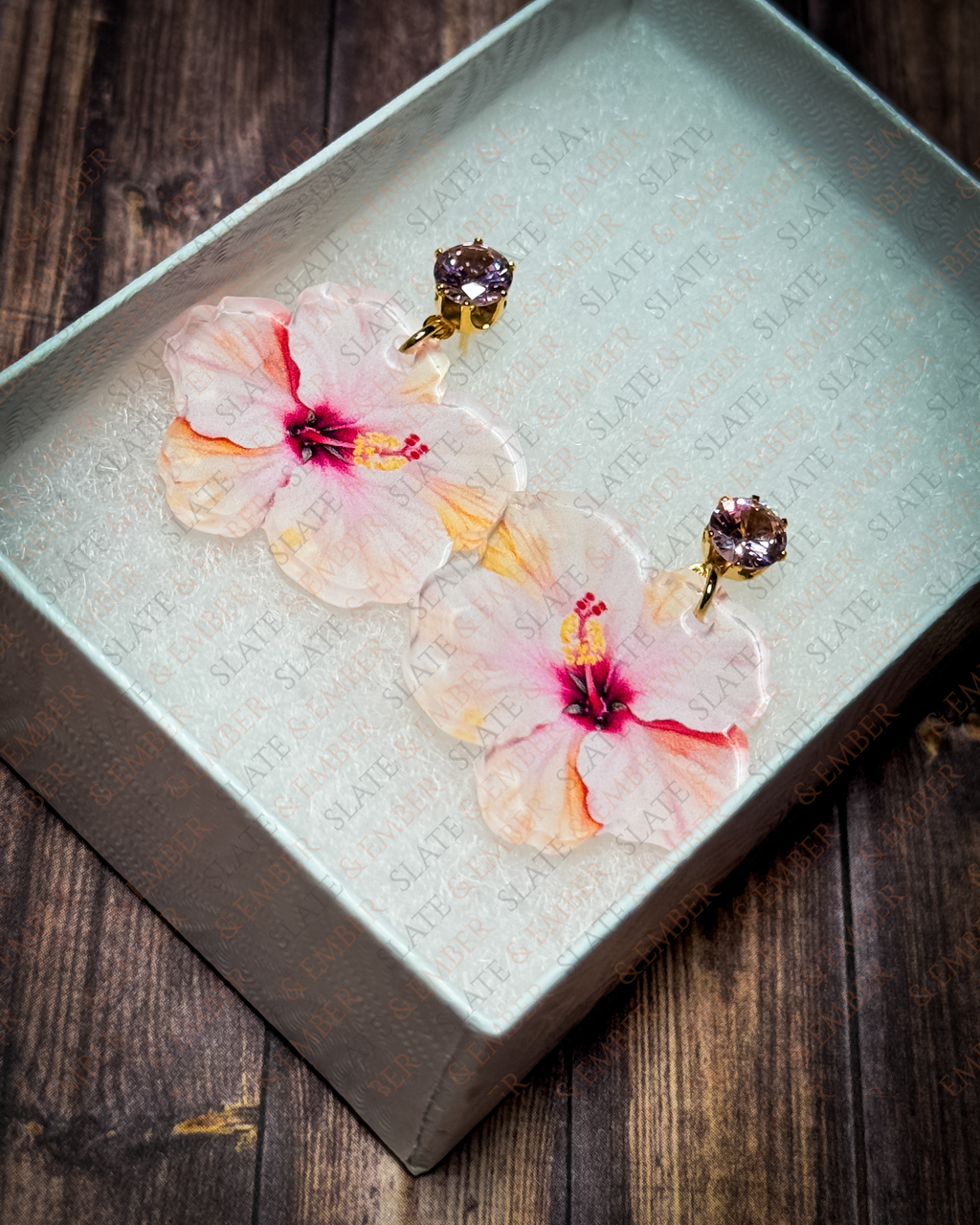 Pink watercolor hibiscus acrylic earrings with pink crystal gold stud tops displayed inside a premium white Slate & Ember jewelry gift box.