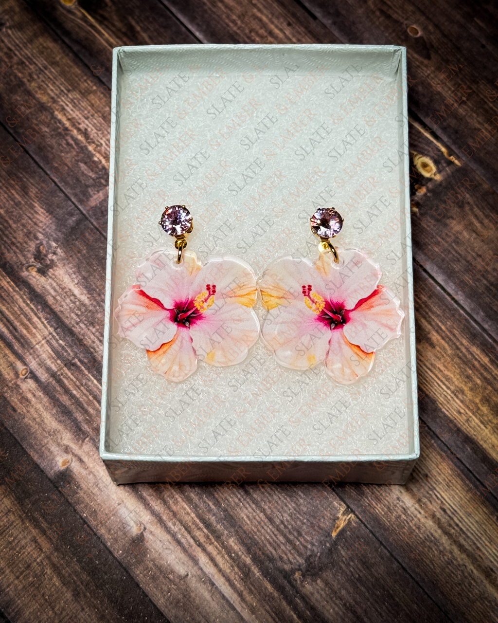 Pink watercolor hibiscus acrylic earrings with pink crystal gold stud tops displayed inside a premium white Slate & Ember jewelry gift box.