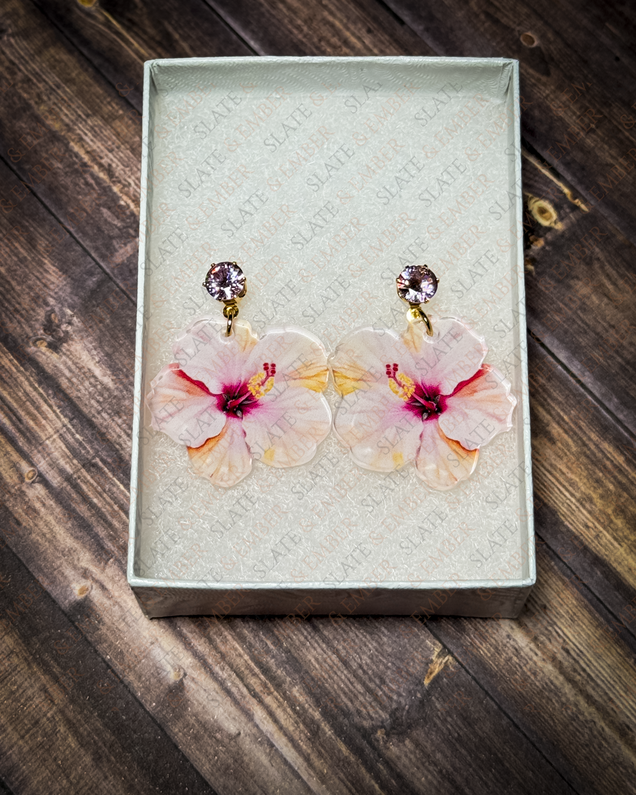 Pink watercolor hibiscus acrylic earrings with pink crystal gold stud tops displayed inside a premium white Slate & Ember jewelry gift box.