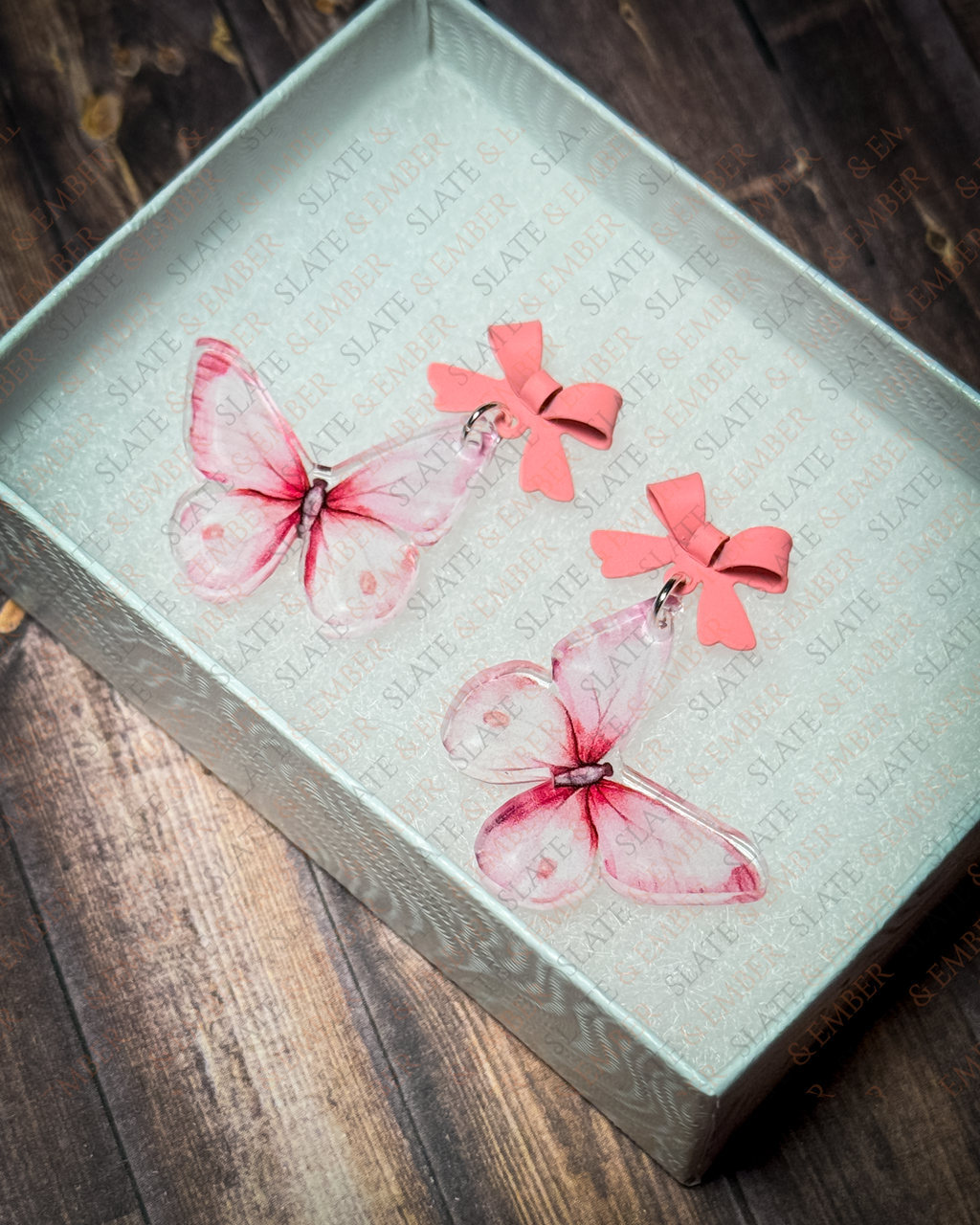 Pink watercolor butterfly acrylic earrings with pink bow stud tops displayed inside a premium white Slate & Ember jewelry gift box.