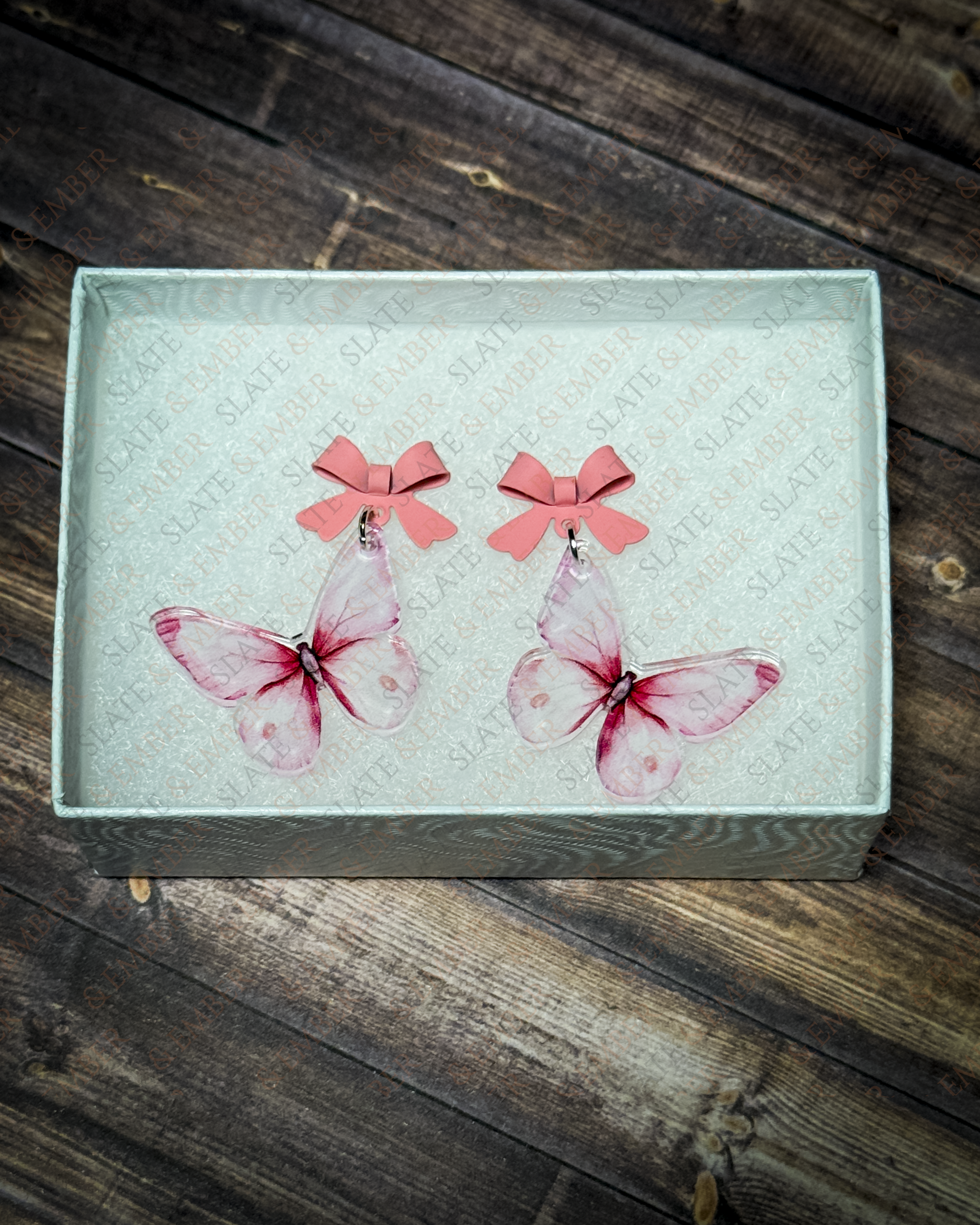 Pink watercolor butterfly acrylic earrings with pink bow stud tops displayed inside a premium white Slate & Ember jewelry gift box.