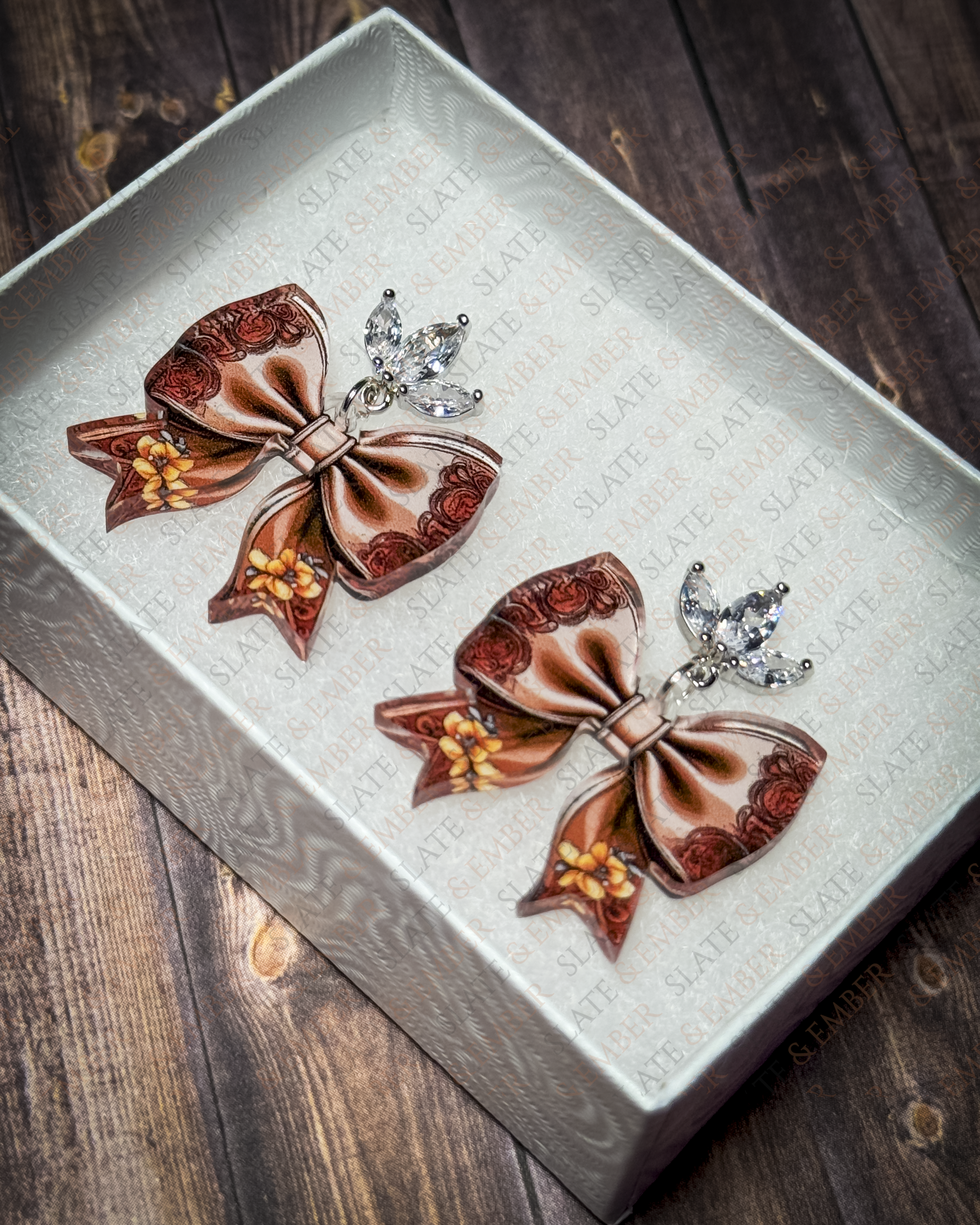 Brown western floral bow acrylic earrings with rose and golden flower details and crystal leaf stud tops displayed inside a white jewelry gift box.