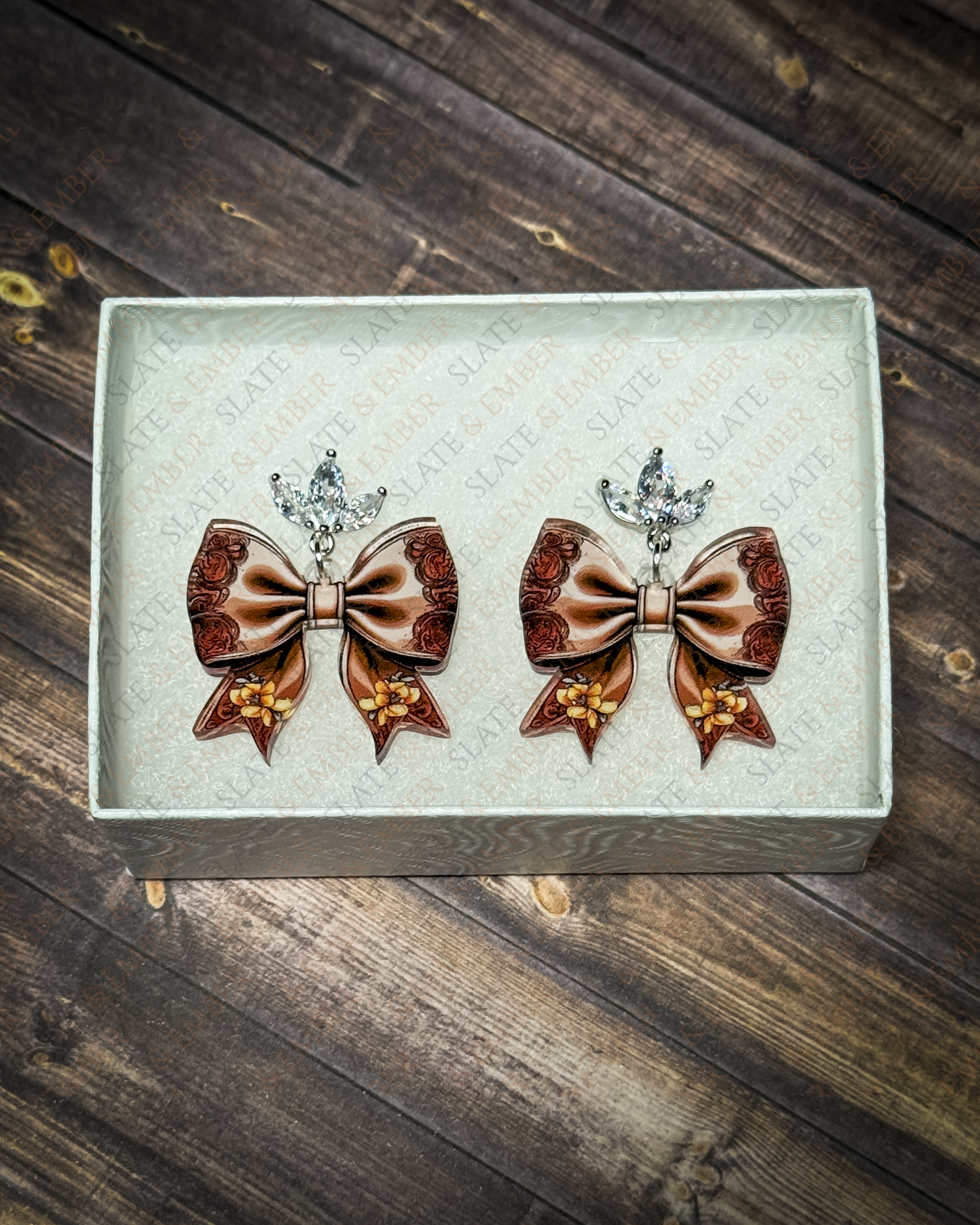 Brown western floral bow acrylic earrings with rose and golden flower details and crystal leaf stud tops displayed inside a white jewelry gift box.